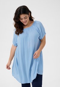 Tunique bleu clair à manches courtes avec un col froncé et une coupe décontractée, réalisée dans un tissu texturé, associée à un jean en denim foncé.