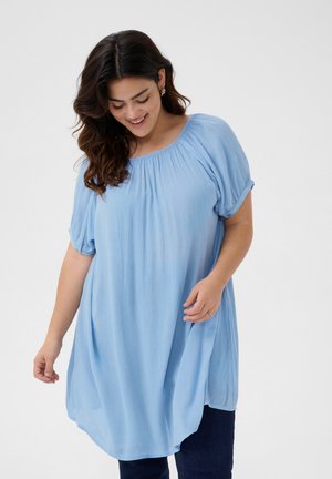 Tunique bleu clair à manches courtes avec un col froncé et une coupe décontractée, réalisée dans un tissu texturé, associée à un jean en denim foncé.
