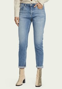 Jeans de mezclilla de lavado claro con diseño de pierna recta, con puños enrollados, cinco bolsillos y cierre de cremallera. Llevados con botas de tobillo de serpiente en color beige.
