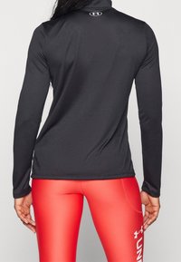 Schwarzes, langärmeliges Sportoberteil mit hohem Kragen und glatter Textur, kombiniert mit enganliegenden roten Leggings, zeigt das Markenlogo auf der Rückseite.