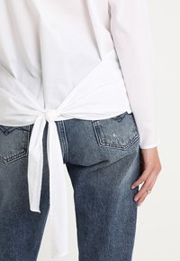 Camicia bianca a maniche lunghe con dettaglio nodo sul retro, abbinata a jeans in denim blu a vita alta. Tessuto morbido, vestibilità casual.