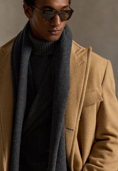 Teveszínű gyapjúkabát bevágott hajtókákkal, sötétszürke sállal és fekete garbóval párosítva. Kerek napszemüveget visel, sima textúrájú.