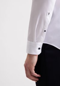 Camicia bianca con maniche lunghe, dotata di polsini con bottoni. Dettaglio ricamato discreto sul polsino. Tessuto liscio e leggero.