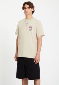 Beige cotton T-shirt with short sleeves and a front graphic featuring a diamond shape with flames and text. Paired with black shorts.