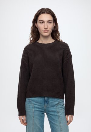 Jeune personne aux longs cheveux bruns portant un pull en maille brun foncé et un jean bleu clair, debout devant un fond clair uni.