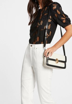 Woman wearing a black lace short-sleeve top, white high-waisted pants, and holding a small white and black crossbody bag with gold clasp.