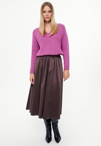 Roze gebreide trui met een V-hals, gecombineerd met een bruine geplooide rok. Model draagt zwarte knielange haklaarzen.