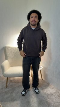 Kohaga lokkis juustega mees, kes kannab tumedat kapuutsi, musti teksapükse ja tenniseid, seisab toas koos valge tekstuurse tooli ja ümmarguse kõrvallauaga.