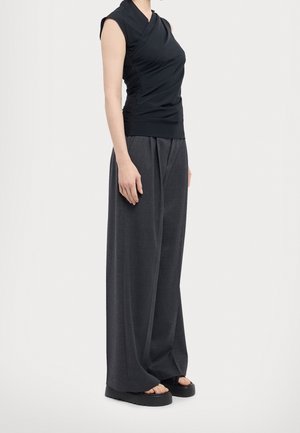 Femme portant un haut noir sans manches froncé, un pantalon gris à jambes larges et des sandales plateformes noires, debout sur un fond unicolore clair.