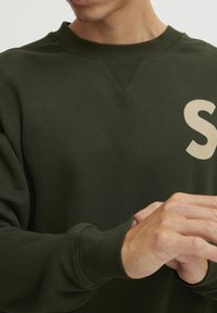 Mørkegrønn sweatshirt med rund hals, ribbede mansjetter og en beige "S"-logo på brystet. Glatt tekstur, minimalistisk design.