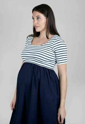 Femme enceinte aux longs cheveux bruns portant une jupe marine et un haut blanc à rayures horizontales noires, regardant légèrement vers la gauche.