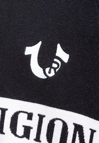 Un material textil negru, cu un logo alb proeminent care seamănă cu un număr "1" întors și un șarpe stilizat, inscripționat cu cuvântul "RELIGION" în litere îndrăznețe.