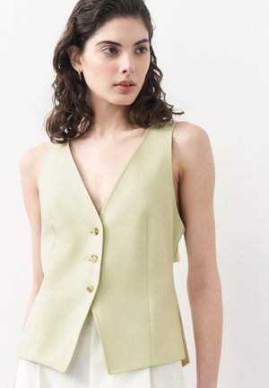 Ženska s temnimi kodrastimi lasmi, oblečena v brezrokaven bledo zelen button-up telovnik in bele hlače, rahlo obrnjena desno na belem ozadju.