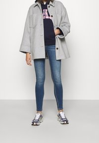 Cappotto oversize grigio in lana con grandi tasche, felpa con cappuccio blu navy con scritte rosa, jeans skinny blu e sneakers multicolori.