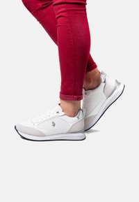 White athletic shoes with perforated design, gray accents, and silver details. Rubber sole and laces. Worn with fitted red pants.