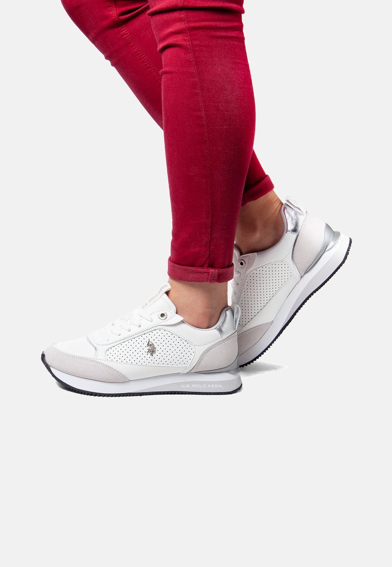 White athletic shoes with perforated design, gray accents, and silver details. Rubber sole and laces. Worn with fitted red pants.