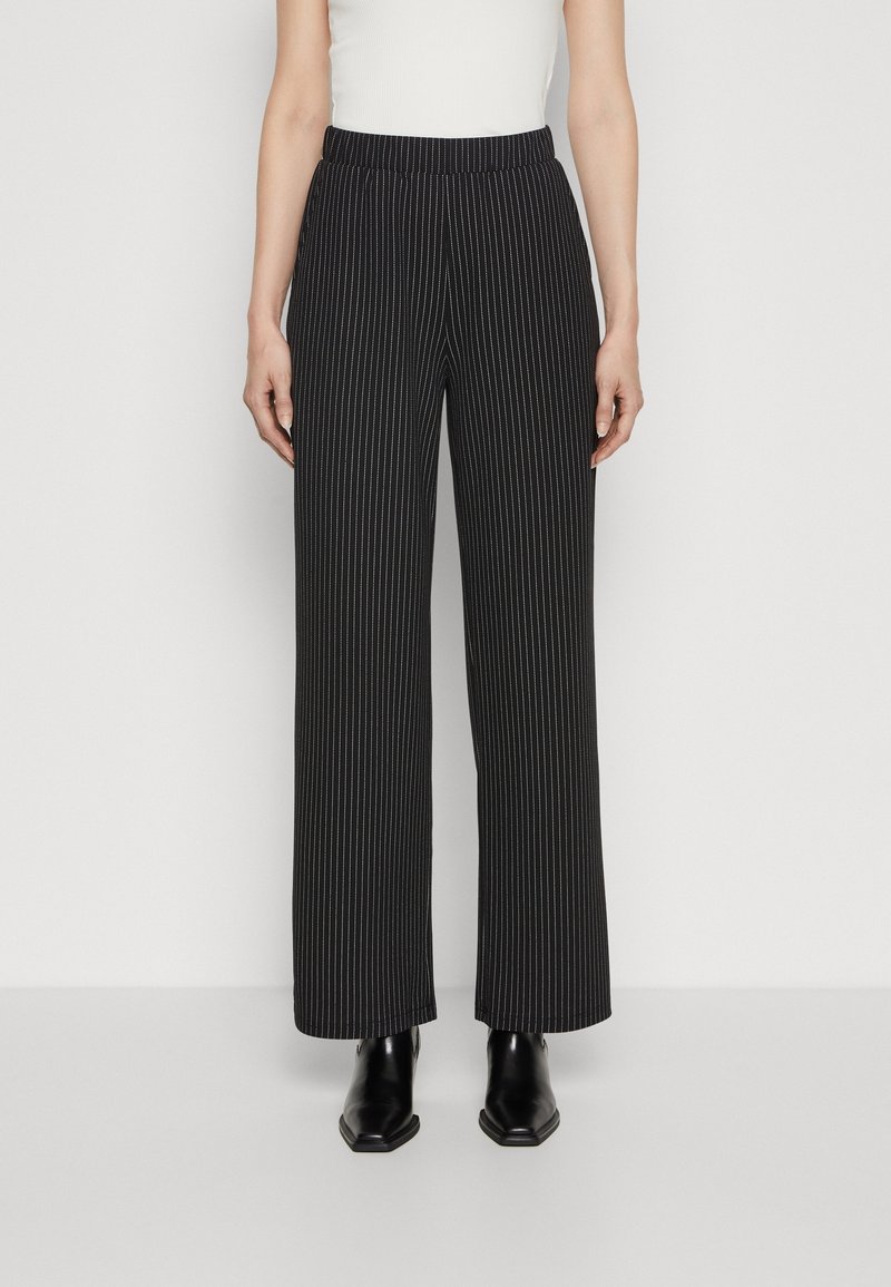Femme portant un pantalon large à fines rayures noires, un haut blanc sans manches et des bottines noires à bout carré, debout devant un fond blanc.