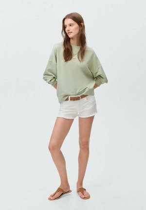 Felpa oversized verde chiaro, pantaloni corti bianchi sfilacciati, cintura in pelle marrone e infradito color sabbia. Design semplice con vestibilità casual e texture morbida.