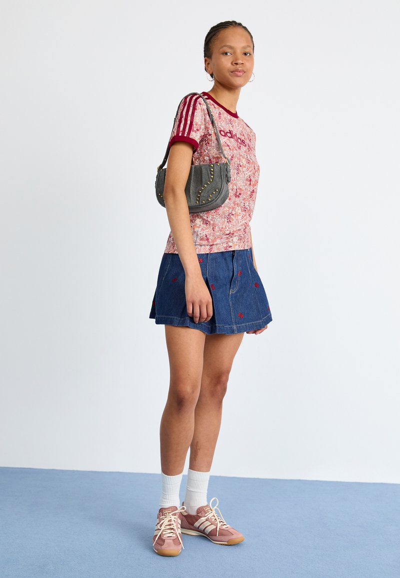 T-shirt à imprimé floral rouge avec des accents bordeaux, jupe en denim bleu avec broderie rouge, sac à main gris orné de clous et chaussures de sport roses avec des chaussettes blanches.