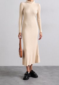 Abito beige a maglia a coste con scollatura ondulata, maniche lunghe e taglio a metà lunghezza. Accompagnato da una piccola borsa marrone e scarpe nere.