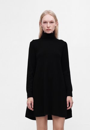 Femme blonde debout, portant une robe noire unie à manches longues et col roulé, expression neutre, fond blanc.