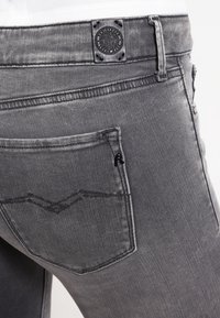 Jeans de mezclilla gris con un bolsillo trasero que presenta un diseño de costura en forma de V y una etiqueta de la marca cerca de la cinturilla. La tela tiene una textura suave.
