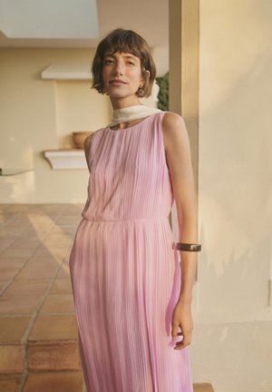 Femme aux cheveux courts et bruns portant une robe plissée rose sans manches et une écharpe blanche, debout à l’intérieur près d’un pilier beige.