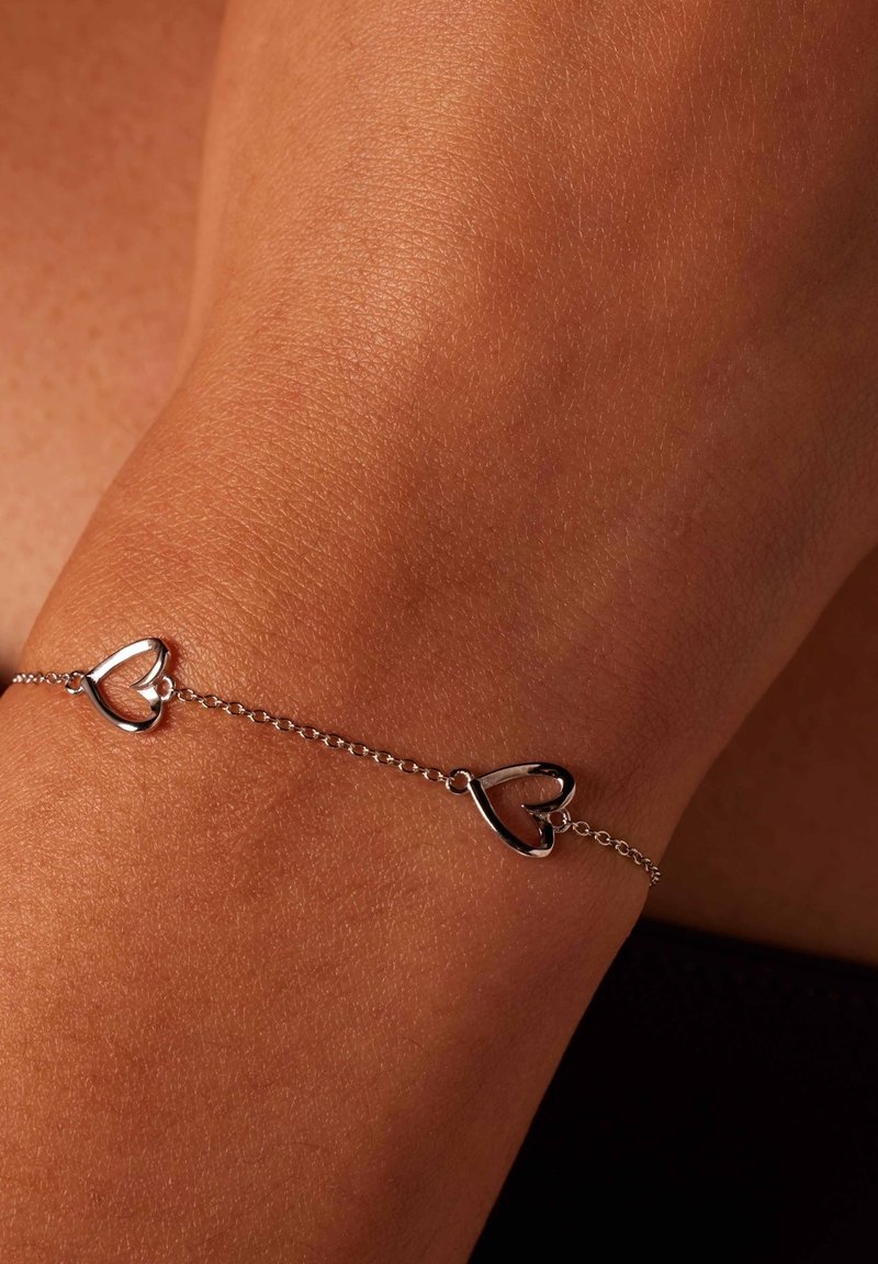 Zilveren kettingarmband met twee hartvormige accenten, tegen een gebruinde huidtoon. Zachte textuur met een verfijnd ontwerp.