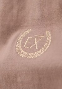 Broderad textil i mjuk taupe med en cirkulär lagerdesign med "EX" i mitten. Slät struktur med subtil lyster.