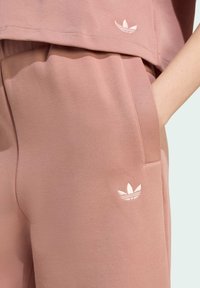 Svijetlo ružičasti sportski komplet koji se sastoji od kratkog topa i hlača s vezicom. Obje komponente imaju mekani izgled i prepoznatljivi izvezeni logo.