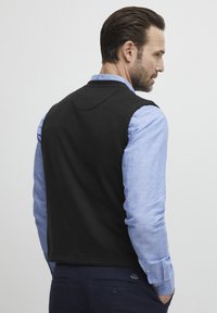 Svart ärmlös tröja över en ljusblå långärmad skjorta. Tröjan har en jämn textur och en rundad halsringning.