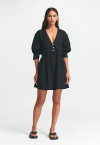 Schwarzes Baumwollkleid mit tiefem V-Ausschnitt, puffigen Ärmeln und Knopfdetails. A-Linie, knielang und mit glatter Textur. Getragen mit schwarzen Sandalen.