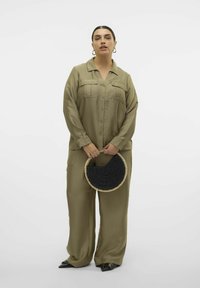 Olivgrön tvådelad outfit bestående av en knapp-up topp med fickor och vida byxor. Håller en rund svart vävd väska med en halmranda.