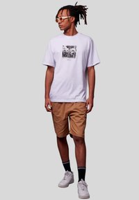 Witte katoenen t-shirt met een grafische print, gecombineerd met beige cargoshorts. Het model draagt witte sneakers en sokken met zwarte strepen.