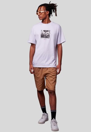 Witte katoenen t-shirt met een grafische print, gecombineerd met beige cargoshorts. Het model draagt witte sneakers en sokken met zwarte strepen.