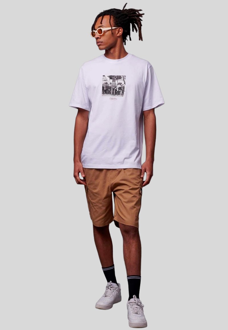 Witte katoenen t-shirt met een grafische print, gecombineerd met beige cargoshorts. Het model draagt witte sneakers en sokken met zwarte strepen.