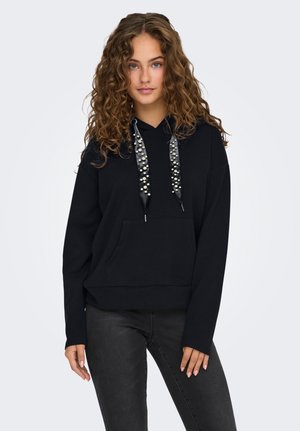 Donna giovane con capelli ricci che indossa una felpa nera con lacci decorati di perle e jeans neri, in piedi su uno sfondo chiaro e uniforme.