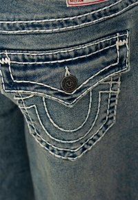 Kieszeń spodni dżinsowych z niebieskim przeszyciem, metalowym guzikiem z logo oraz unikalnym wzorem przeszyć przypominającym kształt litery U, prezentująca wyblakłe, jasnoniebieskie wykończenie.