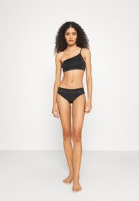 Black one-shoulder bra and matching high-rise bikini set. Smooth texture with logo waistband detailing. Minimalist design and fitted shape.