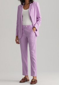 Fialový oblek s přiléhavým blejzrem a kalhotami s rovnými nohavicemi, vyrobený z texturované látky. V kombinaci s bílým tílkem a hnědými mokasíny.