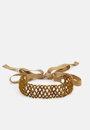 Collier ras de cou doré orné de perles avec un motif en treillis et des liens en tissu beige pour la fixation, présenté sur fond blanc.