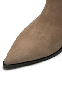 Spitz zulaufender Stiefel aus hellbraunem Suede mit glatter Textur und schwarzer Sohle, der eine taillierte Silhouette präsentiert.