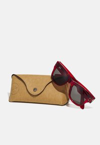 Ochelari de soare roșii cu lentile întunecate, având un cadru geometric îndrăzneț, alături de un toc din suede crem cu închidere cu buton metalic.