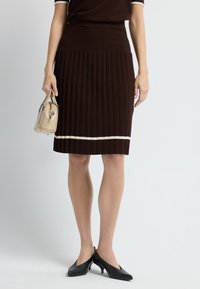 Bruine gebreide geplooide rok met witte horizontale streep aan de zoom, gecombineerd met zwarte hoge hakken en een kleine beige handtas in de hand.