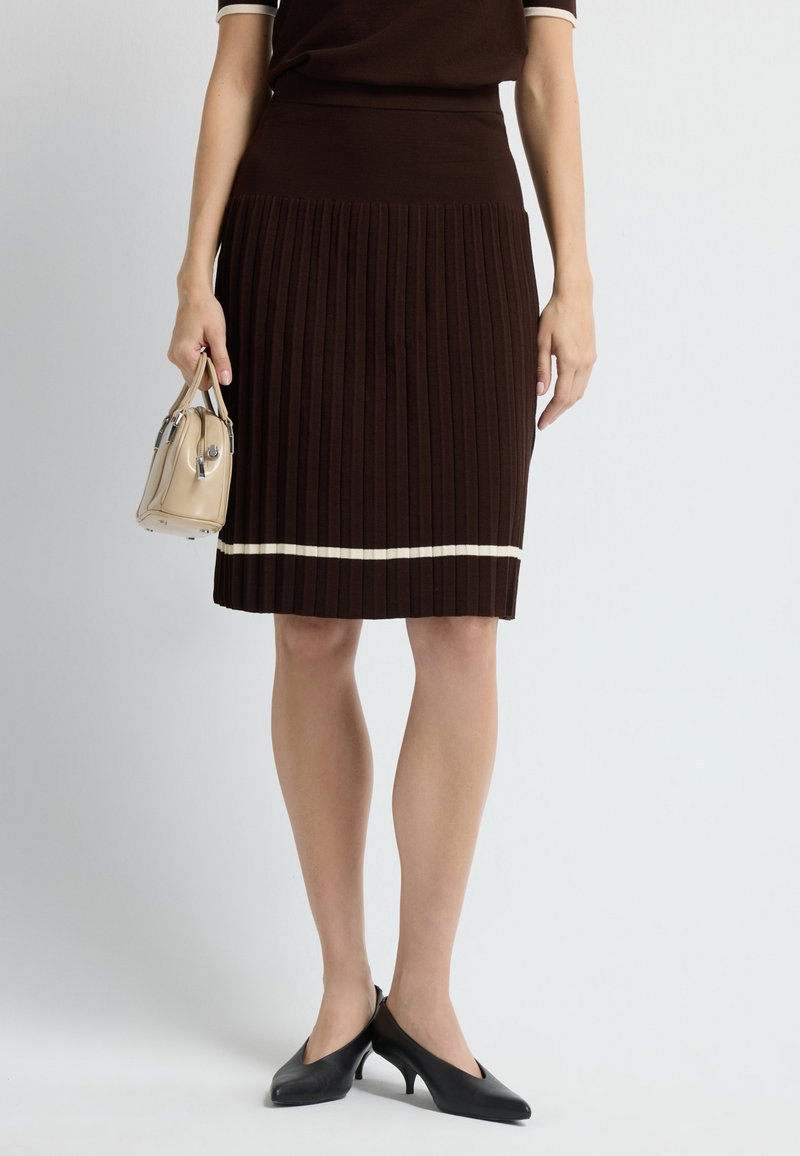 Bruine gebreide geplooide rok met witte horizontale streep aan de zoom, gecombineerd met zwarte hoge hakken en een kleine beige handtas in de hand.