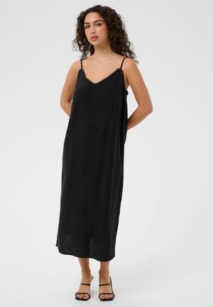 Kobieta o kręconych włosach, ubrana w luźną czarną sukienkę midi na cienkich ramiączkach i czarne sandały z odkrytymi palcami, stojąca na białym tle.