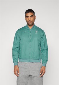 Lysegrøn bomberjakke med lynlås, ribbede manchetter og bund. Har et Liverpool FC-logo på brystet og Nike-branding på siden.