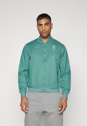 Lysegrønn bomberjakke med glidelås, ribbestrikkede mansjetter og hem. Har Liverpool FC-logo på brystet og Nike-branding på siden.