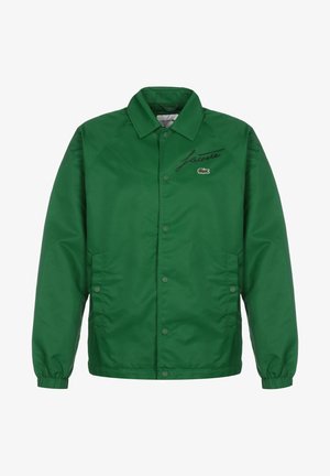 Grøn Lacoste-jakke med knapper, elastiske manchetter, to sidelommer og sort scriptlogo med krokodilleemblem på brystet.