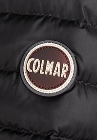 Svart puffjacka i tyg med cirkulär brun läderpatch som bär den präglade texten "COLMAR" i krämfärg. Har en kviltad textur.